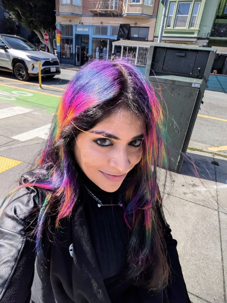 Outdoor front view of a woman in a black jacket with rainbow pixelated hair.