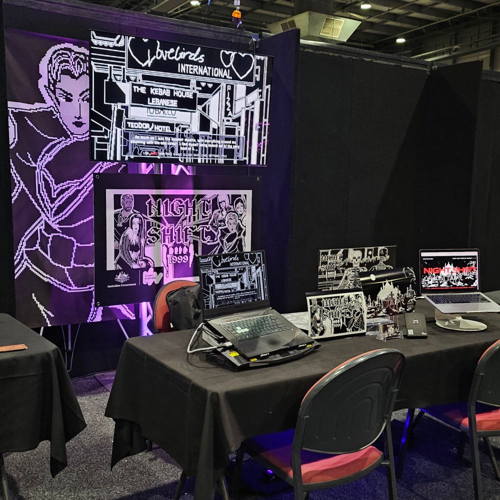 A booth at a convention with two laptops and a TV displaying footage of the visual novel "Night Shift: 1999"