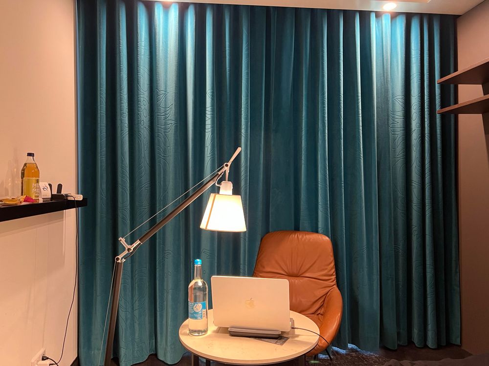 Ein Ledersessel mit einer sehr tief eingestellten Stehlampe vor einem Blauen Vorhang. Ein silbernes Macbook steht auf einem niedrigen Tisch, dem Stuhl zugewandt.