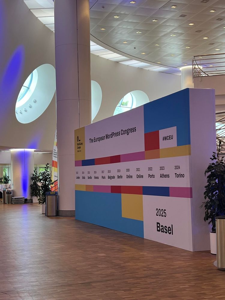 A printed wall with years and locations of previous WordCamp Europes in a big hall.