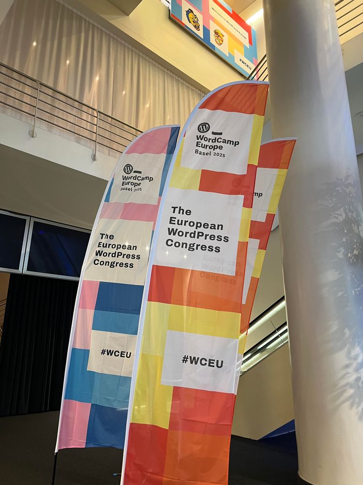 Tall beach flags in a hallway. Vibrant orange, blue and pink pattern. The flags show the WordCamp Europe logo up top, „The European WordPress Congress“ in the middle and the hashtag #WCEU at the bottom