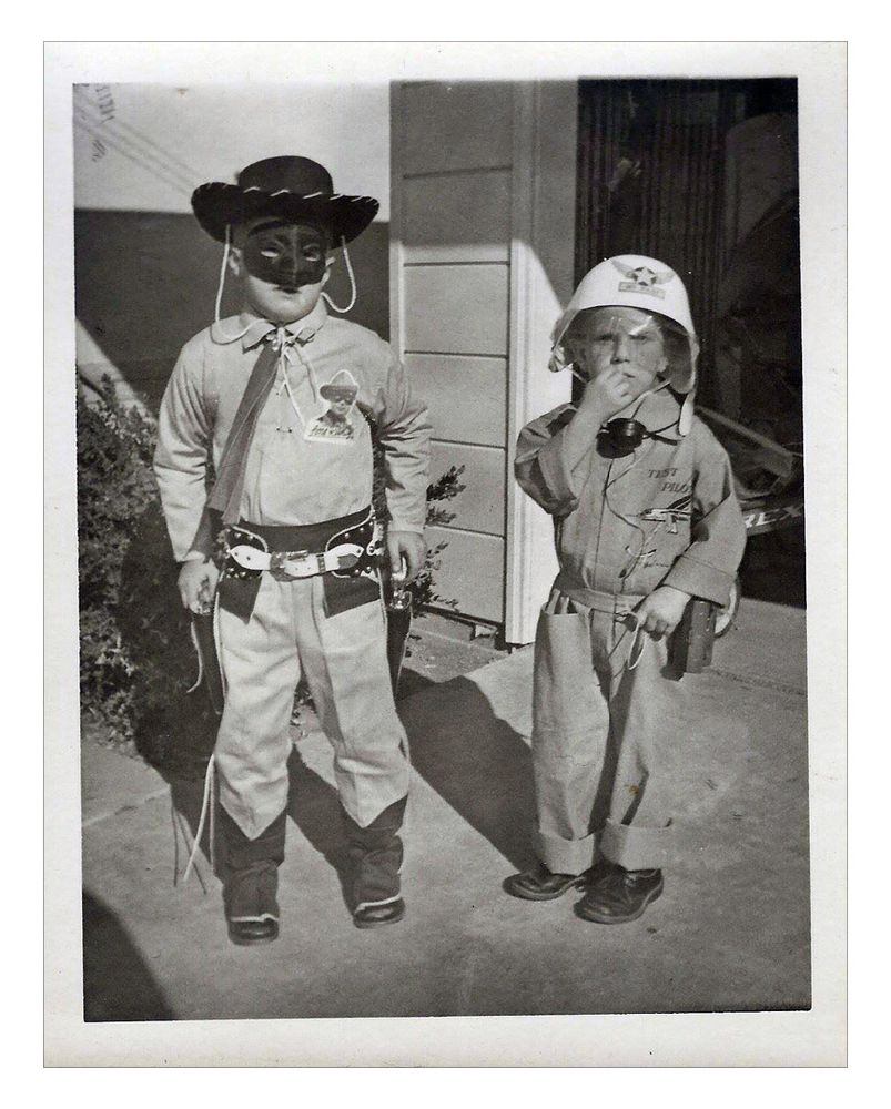 A vintage black and white photograph of two young boys, one dressed as the Lone Ranger, the other in a test pilot costume, both standing on a driveway in front of an open garage.