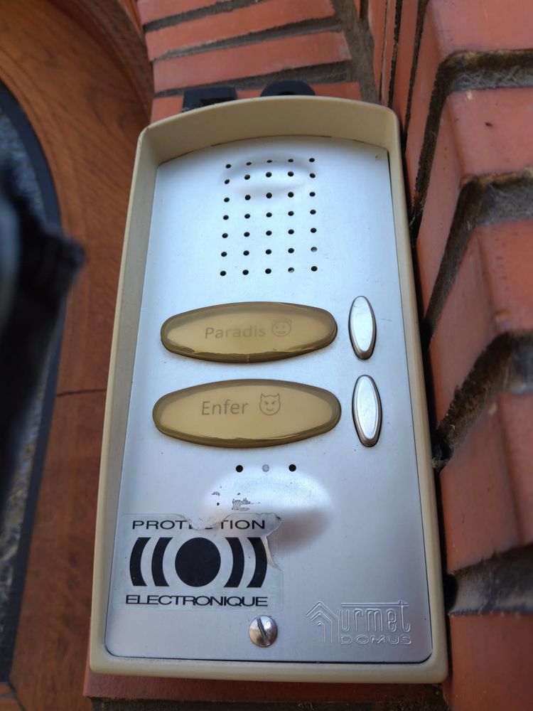 Gros plan d'un Interphone à deux boutons, l'un indiquant paradis, celui du dessous enfer, chacun avec un smiley correspondant. Il est posé sur un mur de briques rouges, près d'une porte en bois arrondie et vitrée 