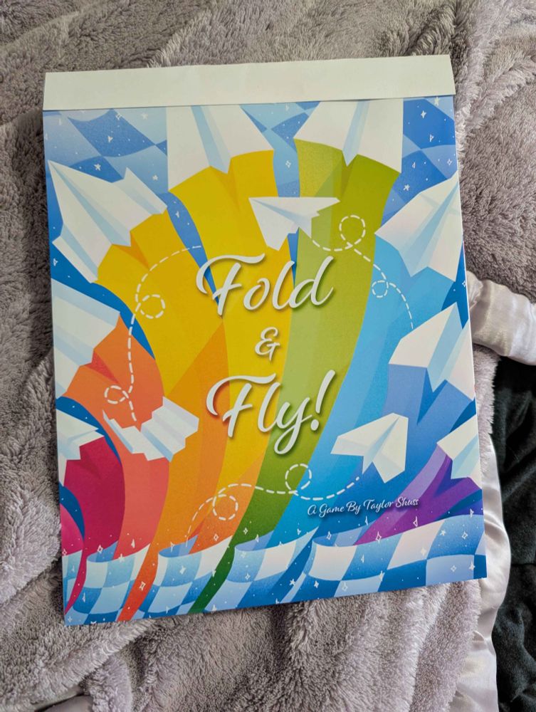 A picture of my IGNM game Fold & Fly!, assembled with the vibrant cover on display. It is on a bed with a blanket below it. The blanket looks incredibly soft & inviting.