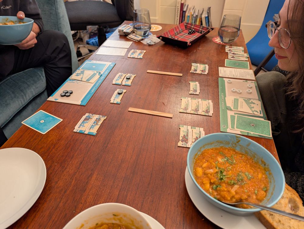 A board game being played on a coffee table. Cardboard ranks of soldiers face off in battle lines. Also there are bowls of bean stew nearby.