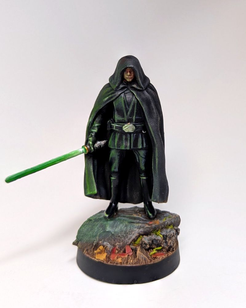 A painted miniature of Luke Skywalker stood on a rock in a black outfit with a black cloak and hood. One arm is under the cloak, the other ends out a hand holding a green light saber to his side.