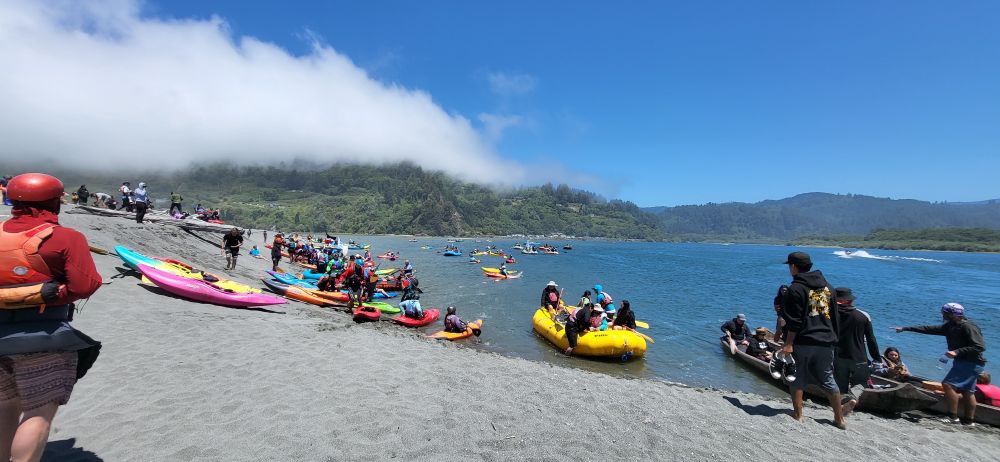 Looking upriver river with green hills now almost fully in view in the background, kayaks & boats lining a sandy shore, a ribbon of fog in the left of the photo - hugging the Ridgeline. The river/estuary a beautiful vibrant blue color. Paddlers are making their way to the boats, some already paddling upriver to the boat dock where food & more fols wait to greet them.