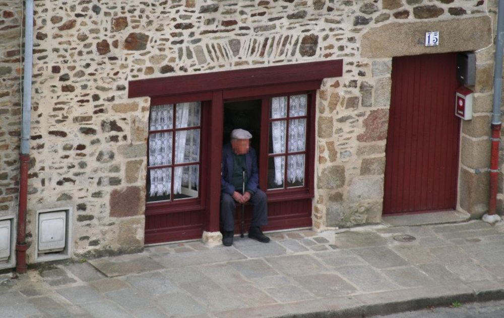 Ein altes bretonisches Steinhaus mit Tür und fast bodentiefem dreifachem Fenster aus rotbraunem Holz; ein alter Mann mit Mütze und Gehstock sitzt im offenen Fenster.