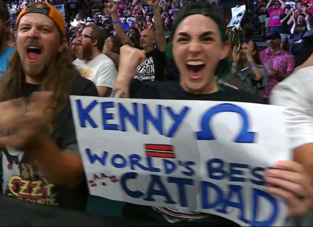 Enthusiastic fan holding a sign that reads "Kenny [Omega symbol] = World's Best Cat Dad"