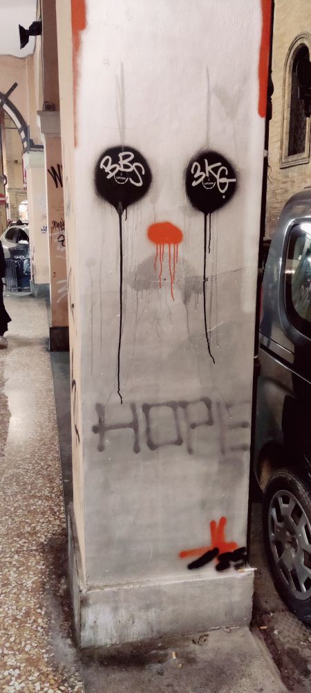 En buegangssøyle hvor 2 svarte, taggede ballonger, en rød rennende sky under og en utvisket grå skygge lager et trist klovnefjes.

Teksten "Hope" er skrevet under.