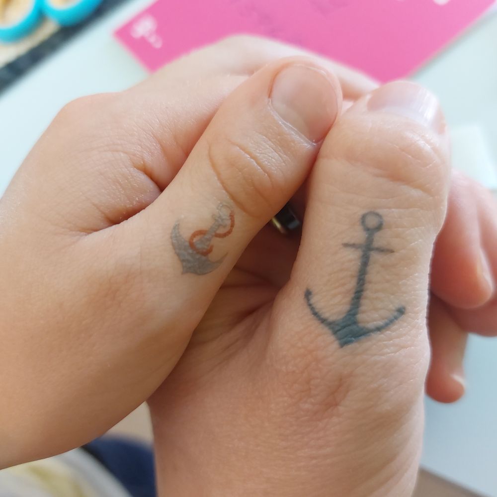 Eine Erwachsenenhand und eine Kinderhand. Beide haben einen Anker auf dem Daumen, eins ist echt und eins ist geklebt. 