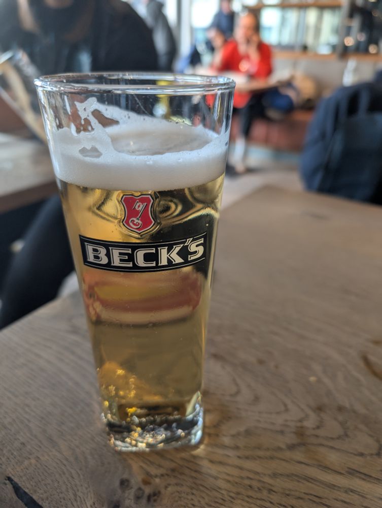 Auf dem Bild steht ein hohes, schlankes Bierglas auf einem Holztisch. Das Glas ist mit hellem Bier gefüllt und trägt das Logo der Marke Beck’s auf der Vorderseite. Oben im Glas befindet sich ein Schaumkranz. Der Hintergrund ist unscharf, aber man erkennt, dass sich das Glas in einer Art Café oder Bar befindet. Einige Personen sitzen oder stehen im Hintergrund, wirken jedoch verschwommen und sind nicht klar erkennbar. Die Atmosphäre scheint entspannt und zwanglos zu sein.