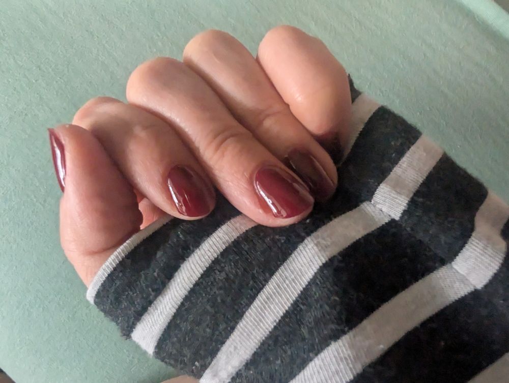 Das Bild zeigt eine Hand, die leicht zur Faust geballt ist. Die Fingernägel sind kurz bis mittellang und mit einem glänzenden dunkelroten Nagelfolien beklebt. Die Person trägt ein langärmliges Kleidungsstück mit schwarz-weißen Querstreifen. Der Hintergrund ist einfarbig in einem hellgrünen Ton gehalten.

