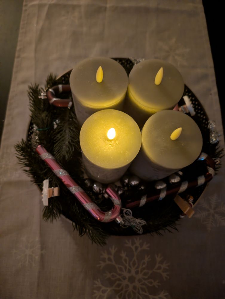 

Auf dem Foto steht ein adventlicher Kranz in der Mitte eines Tisches. Der Kranz ist dekoriert mit künstlichem Tannengrün, glänzenden silbernen Elementen und mehreren Zuckerstangen in Rot-Weiß und Silber. In der Mitte des Kranzes befinden sich vier große, zylindrische LED-Stumpenkerzen in einem warmen Grauton. Eine der Kerzen leuchtet mit einer warmen gelben Flamme, während die anderen drei ausgeschaltet sind. Der Kranz steht auf einer Tischdecke mit dezenten Schneeflockenmustern, was dem Arrangement eine winterliche und festliche Atmosphäre verleiht.