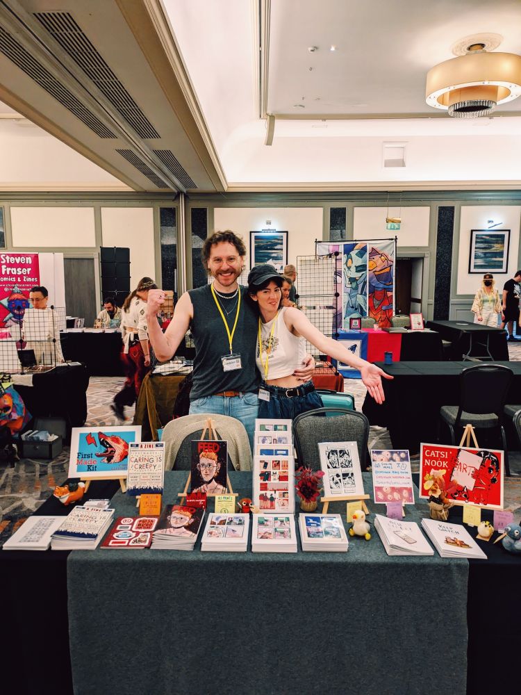 Two dorks selling zines at their convention table. 