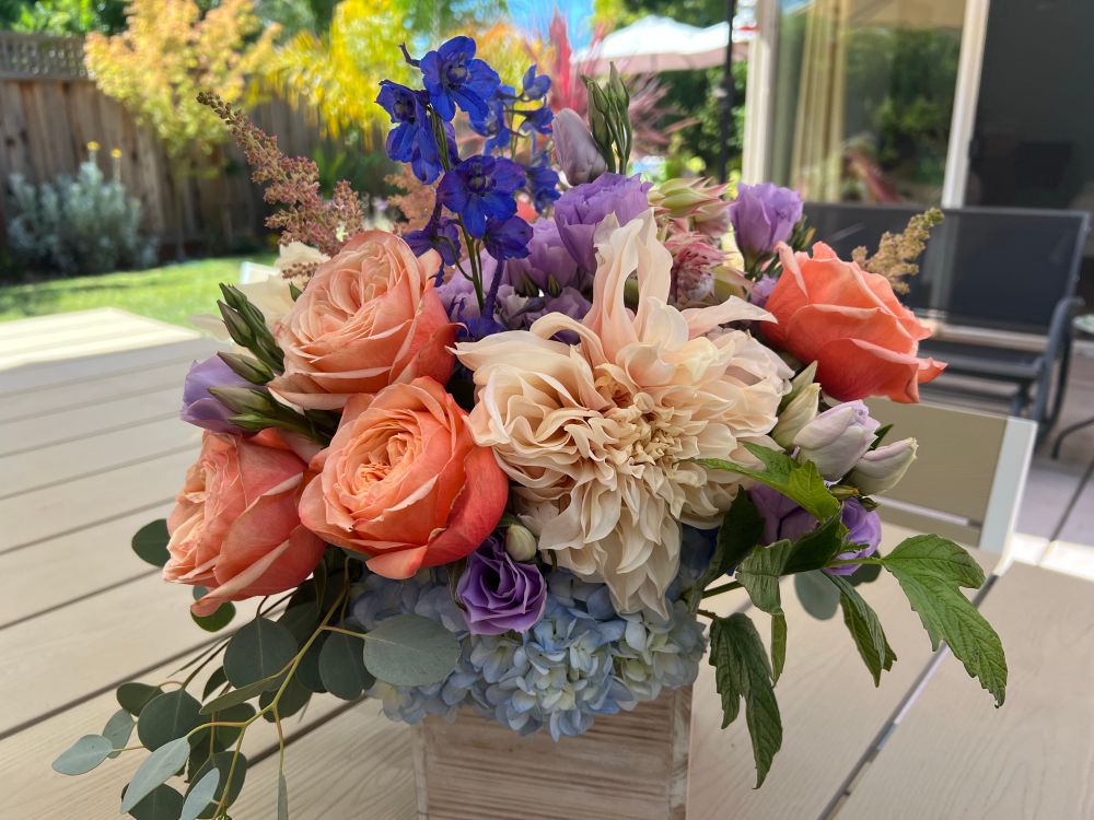 Orange roses, Café Olay, big Dalia, greenery, purple lisanthus, blue Delphinium, all in a bouquet on a grayish tan table.