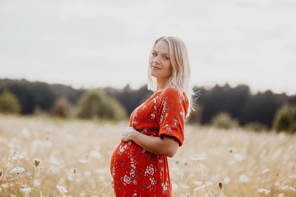 a pregnant blond woman in a red flowered dress standing in a field.