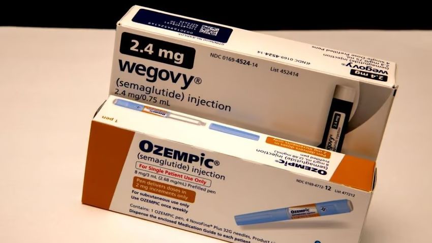 A box of Ozempic in orange and white with a blue drug pen on it. Behind it is a box of Wegovy. The box is white with navy blue writing and a blue and white pen.