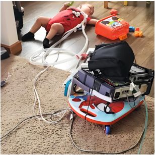 baby is on the floor playing with some blocks. He is lying on his back. He has a ventilator attached to his neck. A ventilator is on a rolling child's toy. Cords are also attached.