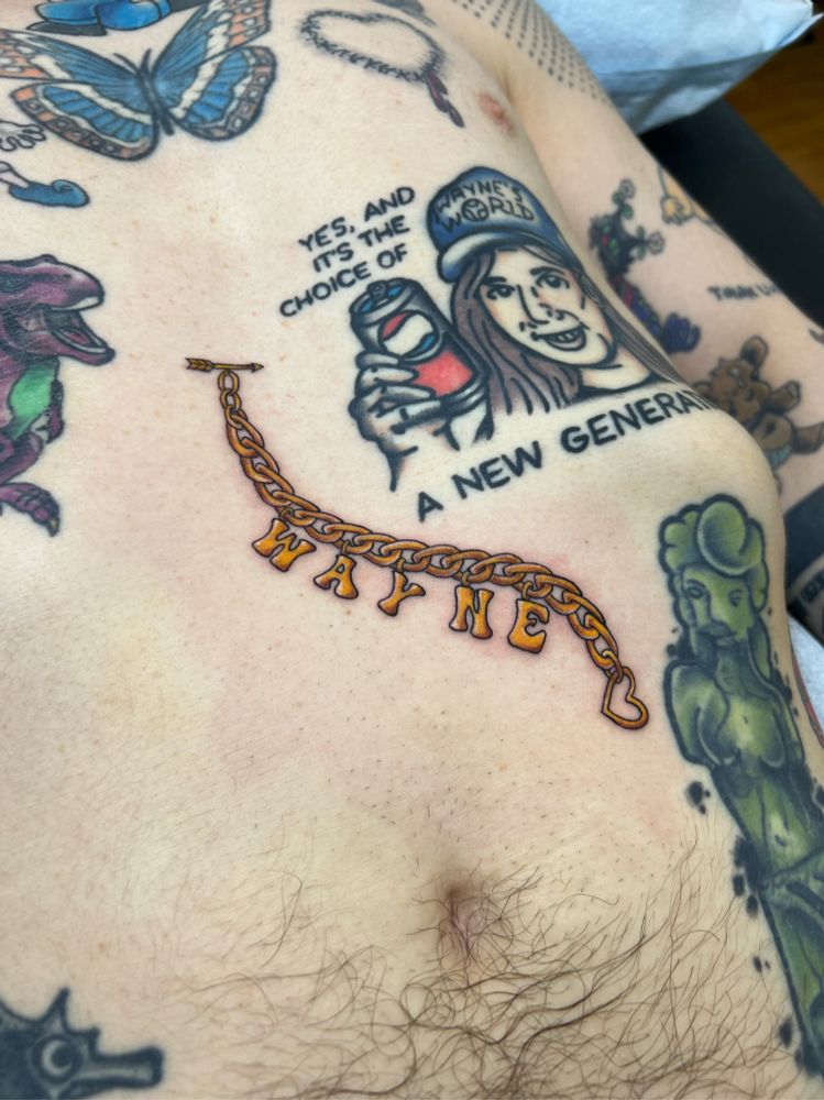 A photo of a fresh tattoo on a persons torso. The tattoo features a gold chain with letter charms spelling out the name Wayne. 