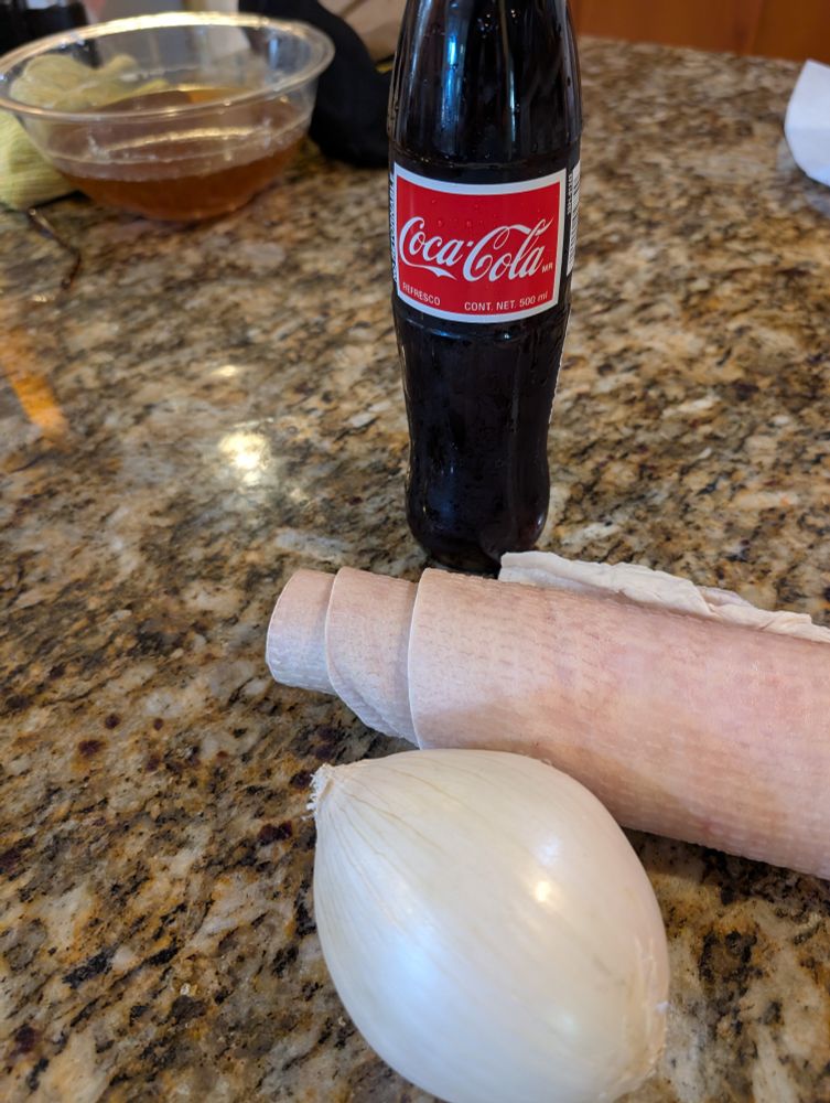 A bottle of Mexican Coca-Cola, a mysterious pale pink rolled object, and a white onion stop a granite countertop 