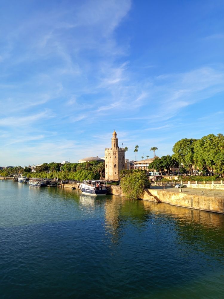 The Torre del Ore, a squat medieval tower on the banks of the Guadalquivir that used to hold all the stolen treasures from the New World. Above a blue sky strafed by clouds. A barge sits at harbour. 