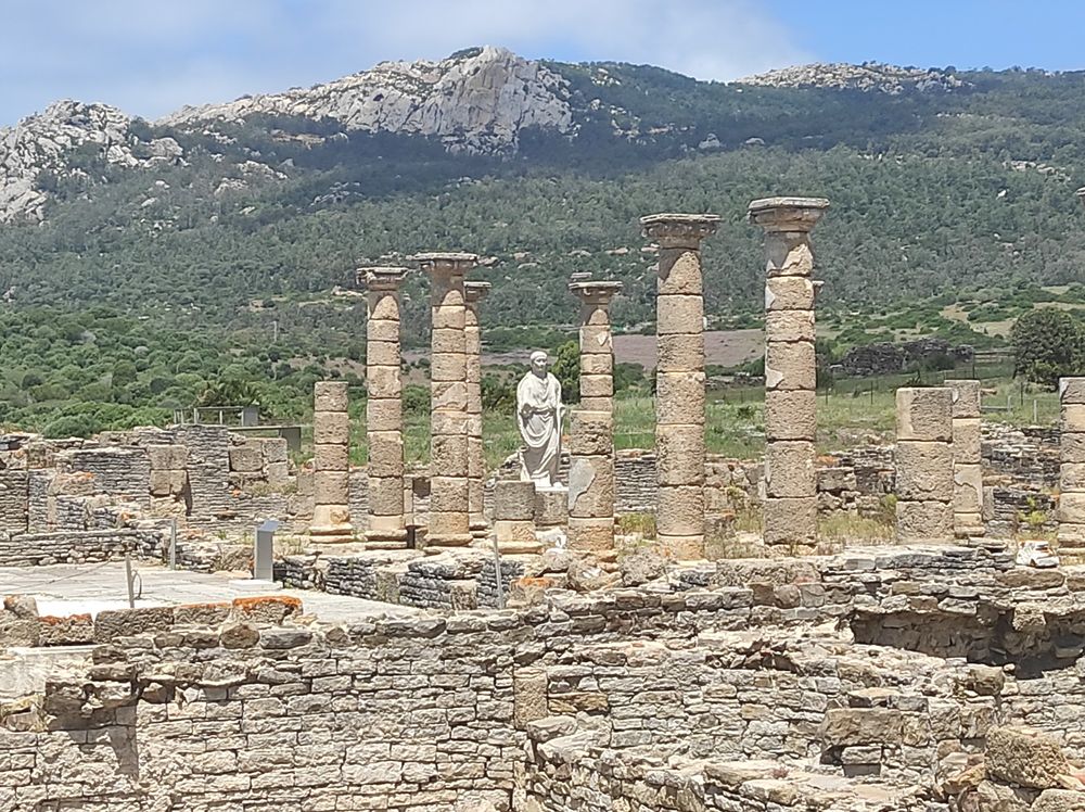 A Roman statue and ruins on the coast of Bolonia, Spain. A grey mountain rises in the background. 