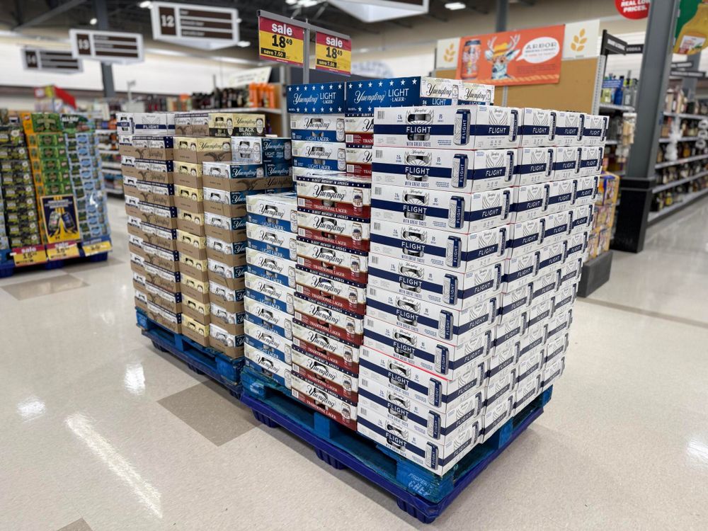A display of beer boxes, including varieties like Yuengling Light, Yuengling Black, and Yuengling Flight, stacked on pallets in a store aisle. A clearance sale sign is visible above the display.