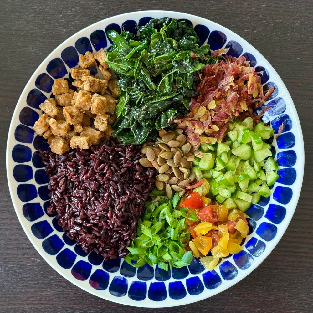 A vibrant bowl of food featuring sections of black rice, massaged kale, diced cucumbers, yellow and red cherry tomatoes, green onions, freshly toasted pumpkin seeds, shredded purple carrots, and air fried tofu. The bowl has a decorative blue pattern against a dark wooden table.