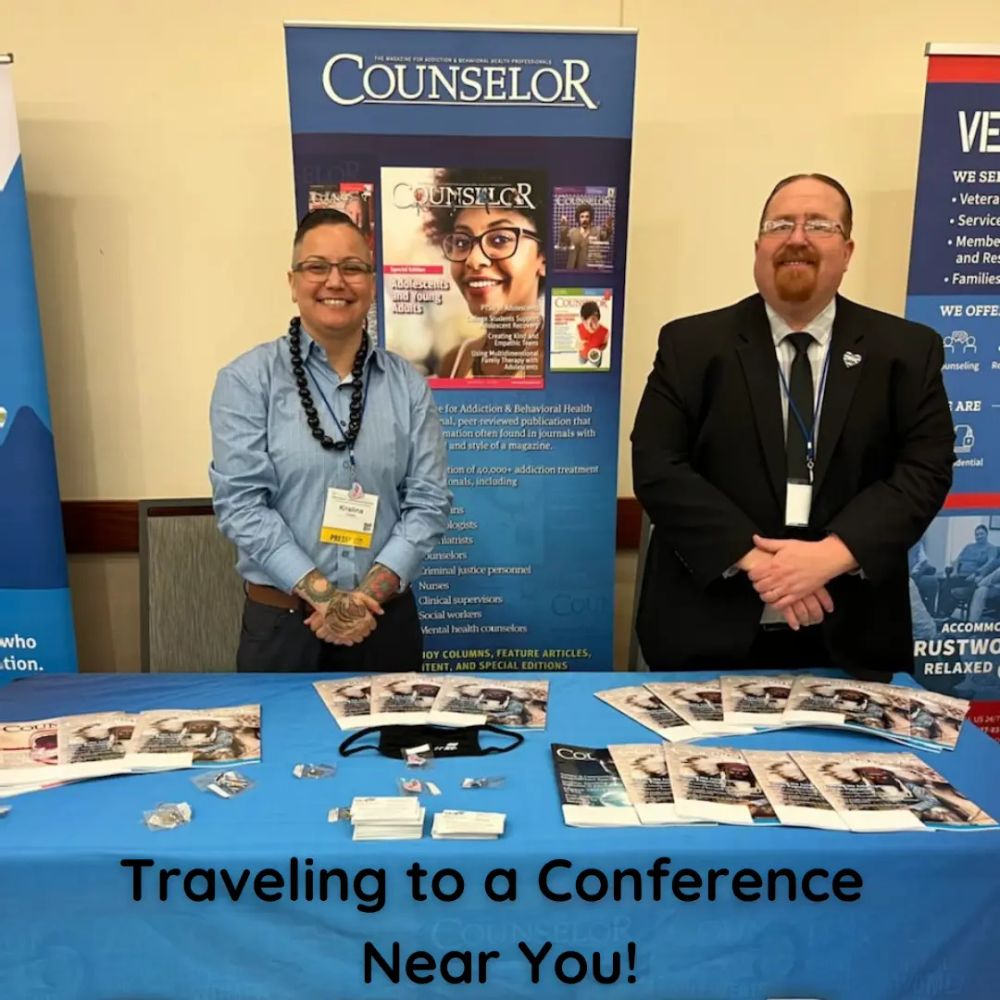 two people standing behind an exhibit table. One is non-binary with short black hair and wearing a long-sleeved blue shirt with a beaded necklace. The other is a white man with red hair and beard, wearing glasses and a black suit. The exhibit table says 'Counselor' and has examples of the magazine.