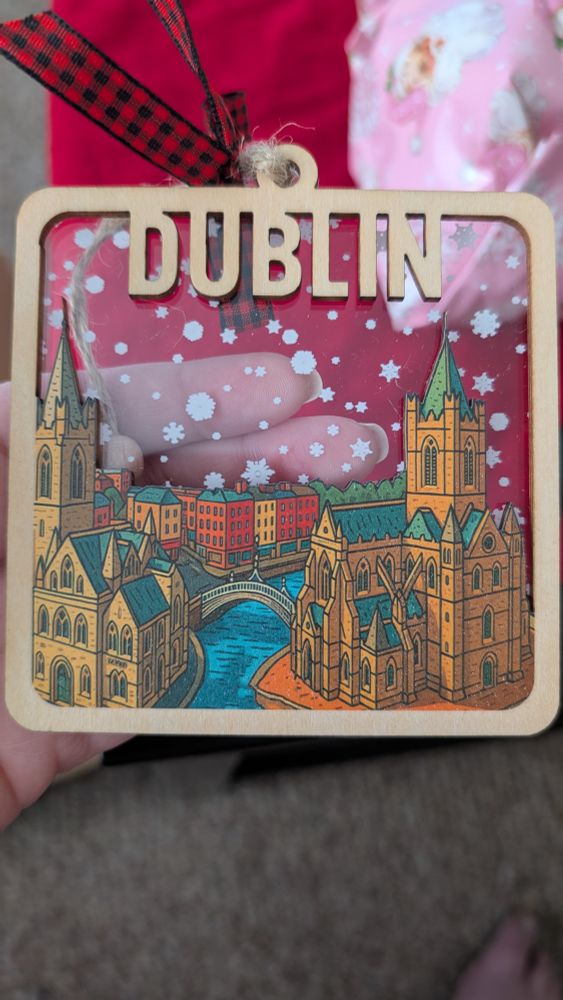 An acrylic and wooden Christmas ornament, made of a panel of acrylic with snowflakes on it fronted by a carved wooden panel of old Dublin in a cartoony style showing St. Patrick's cathedral, Christchurch cathedral, the Happenny Bridge and Temple Bar with the word DUBLIN over it. 