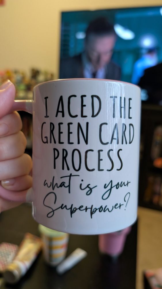 A white mug with a pink handle, that says I aced the green card process. What is your superpower?