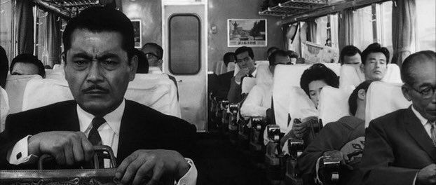 Mustached man gripping his briefcase tightly, face scrunched up in a scowl, as he rides in a filled passenger car. 