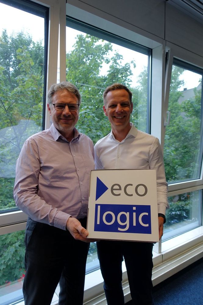 Christoph Heinrich und Kai Niebert vor einem Fenster, im HIntergrund sind Bäume zu sehen. Sie halten einen Würfel mit der Aufschrift "ecologic"