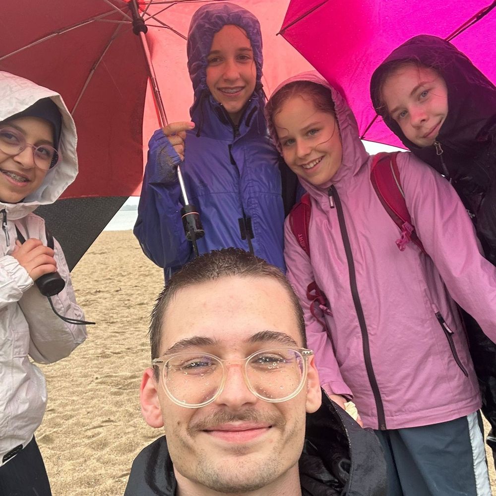 Ein junger Mann macht ein Selfie mit vier Schüler:innen, die unter bunten Regenschirmen am Strand stehen. Alle tragen Regenjacken und lachen in die Kamera.