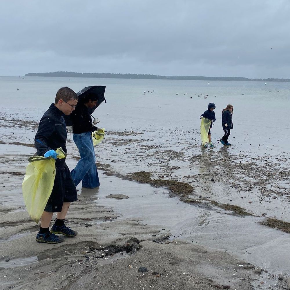 Vier Kinder laufen bei Regen mit gelben Müllsäcken entlang eines grauen Strandes. 