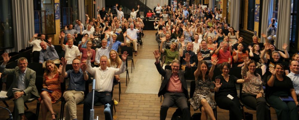 Viele Personen sitzen in einem großen Saal, lachen und winken bzw. recken die Hände empor.
