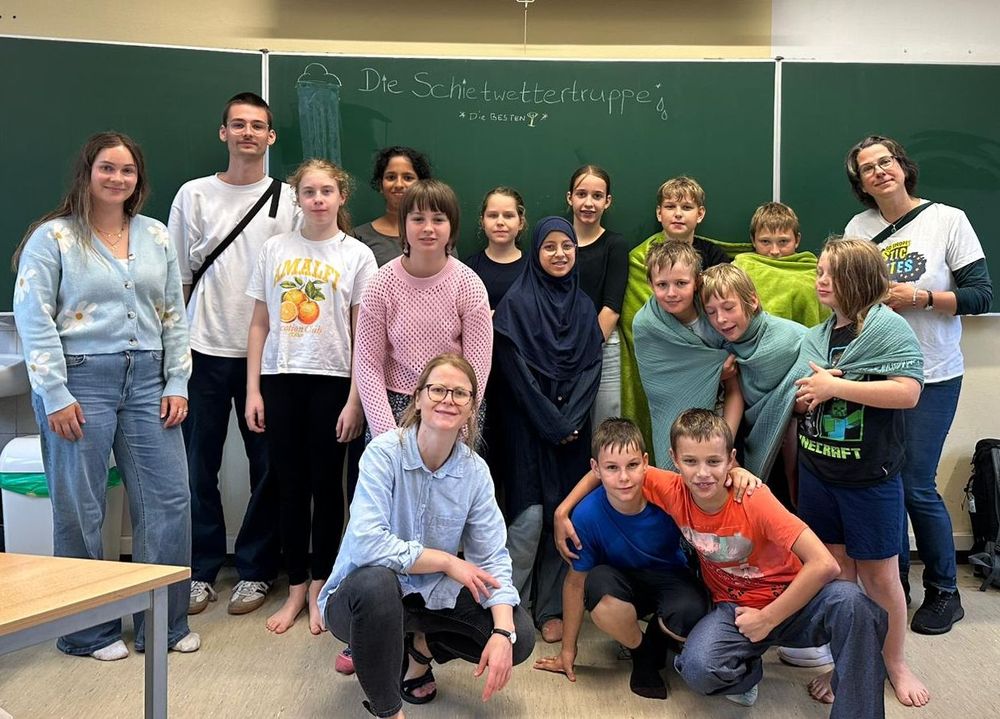 Schülerinnen und Schüler der Klasse 5A sowie zwei Erwachsene stehen vor einer Tafel mit der Aufschrift „Schietwettertruppe“. Einige Kinder sind in Handtücher gewickelt.