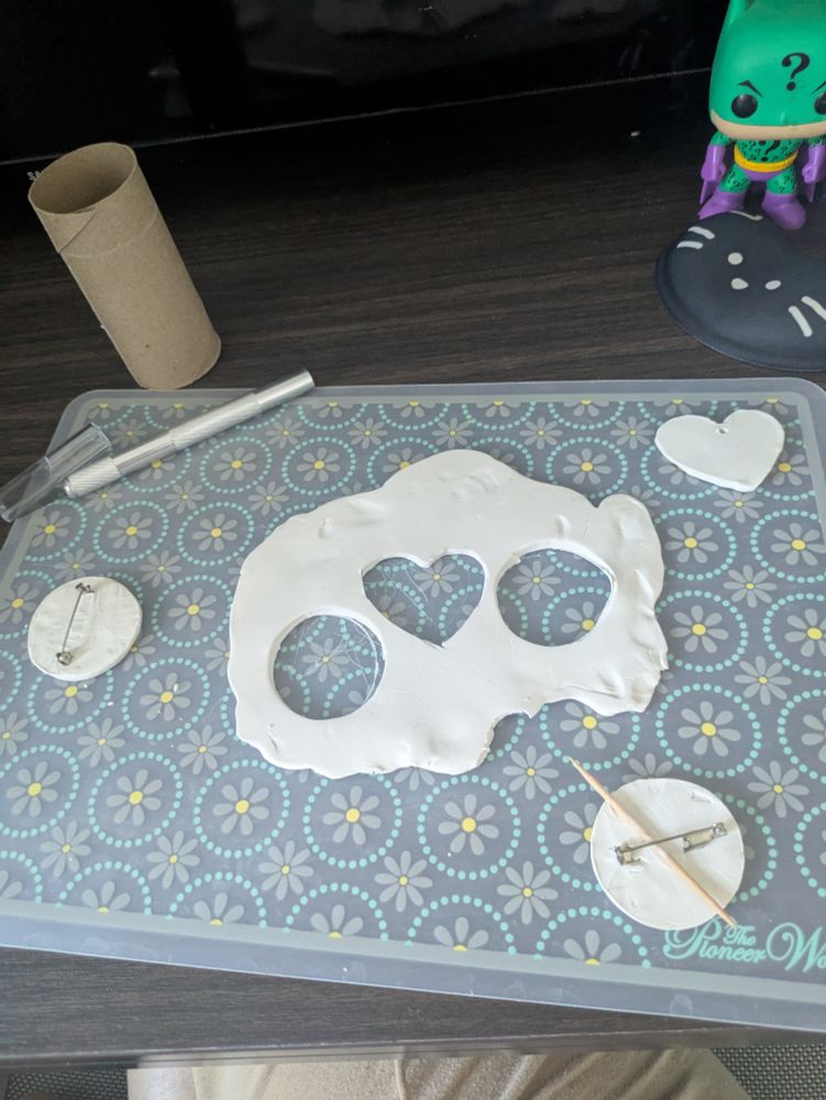 A blue, flowery cutting mat with a mat of clay in the center. The mat has a circle, a heart, and another circle cut out. Around it is the heart with a hole at the top to be a charm, and the two circles with flat-edge pins in it to be button pins. The pin on the bottom right has a toothpick on it, used to flatten the flat edge of the pin in place with clay. The pin on the upper left has an exacto-knife and a cardboard roll next to it that was used for carving the shapes out of the clay mat.

 A cat themed wrist rest and a Funko Pop of Batman in a Riddler themed suit can be seen in the upper right corner