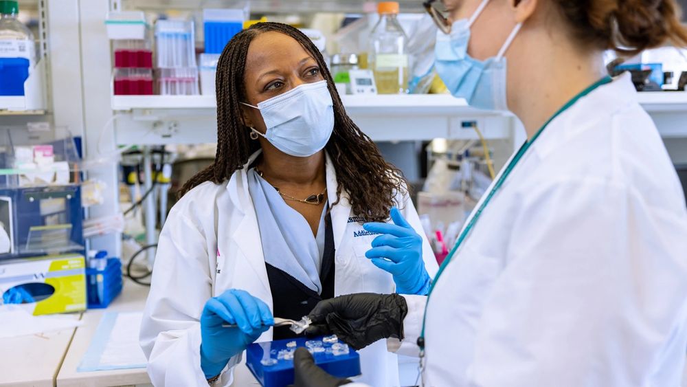 Photo of Dr. Yasmin Hurd and a member of her lab. They are in the lab wearing lab coats, masks, and gloves and are handling lab equipment.