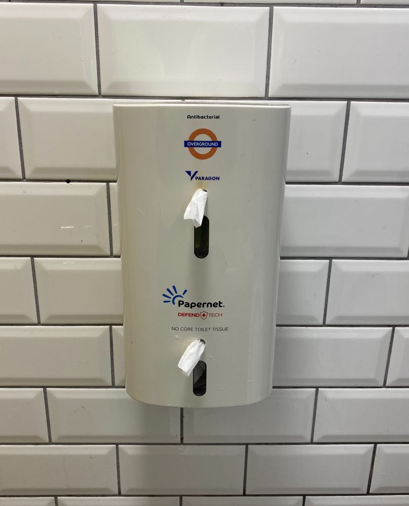 Single-sheet toilet dispenser with small London Overground roundel sticker, on white beveled brick-style tiles.