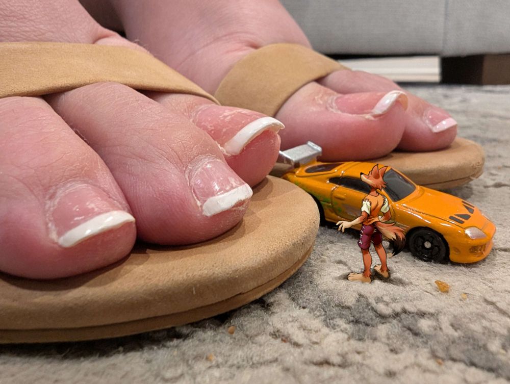 picture close to the ground of Nikki's toes in her open-toe heels, with a small orange toy car between them, and a tiny Alem drawn in standing in front of her feet