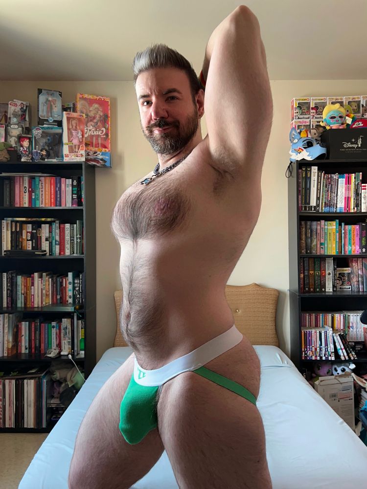 Standing at the end of my bed in a green jockstrap with my arm behind my head 