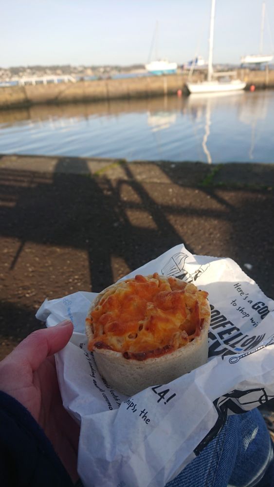I'm holding a macaroni pie on a paper bag. The view is a sunny marina. 