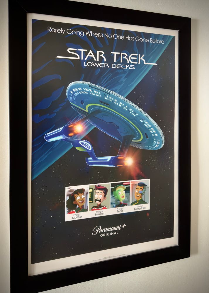 A 27x40 inch poster for the animated series Star Trek: The Lower Decks in a black spotlight hot swap display frame on a white plaster wall giving off Southern California post-war vibes. The poster is by Matt Ferguson and says “Rarely Going Where No One Has Gone Before, Star Trek: Lower Decks”. Below the text, the USS Cerritos, a California class starship, flies towards the viewer. Below the Cerritos are the four main characters of the series. The poster is by Matt Ferguson. It look better than a Kirk sundae with Trip Tucker sprinkles.