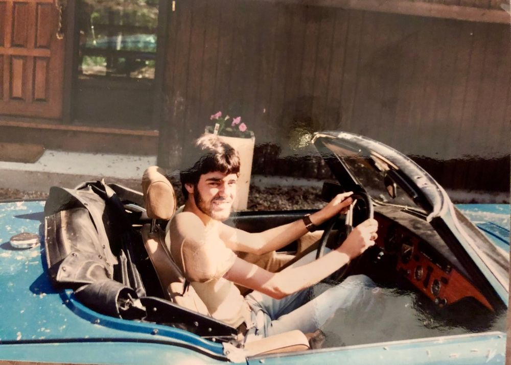 A man in his late teens or early 20s behind the wheel of a blue Triumph Spitfire with the top down. He's wearing blue jeans and a pale orange tee shirt. He has dark brown hair and a full beard. The car is parked in front of a house with dark brown siding and a brown door.