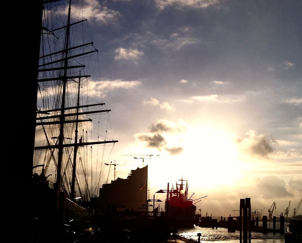 Hafenansicht als schwarze filigrane Silhouette vor leicht bewölktem Himmel mit einer großen Fläche an durchbrechendem Sonnenlicht. Neben den Umrissen von Schiffen und Kränen ist auch die Elbphilharmonie zu erkennen - hier noch vor ihrer Fertigstellung