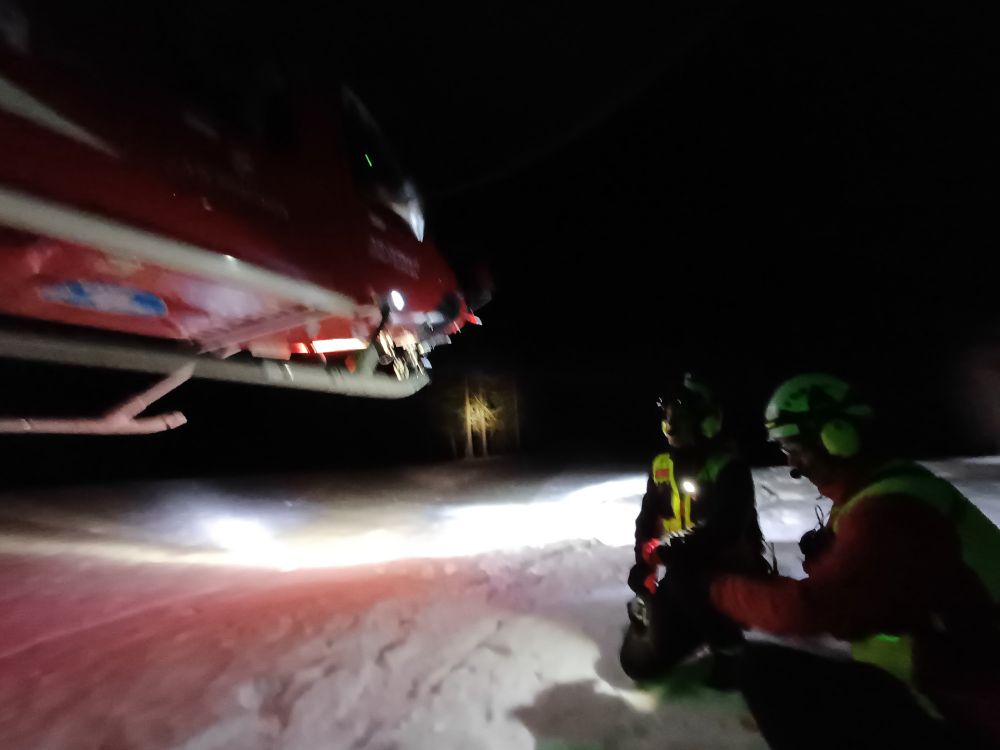A helicopter hovering over two trainees above the snow in the dark.