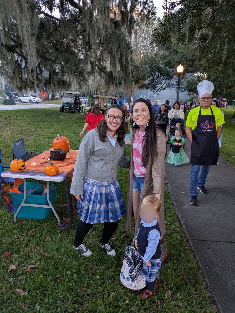 Photo of me standing with a woman and her child. The child and I are both dressed up as Rory Gilmore, both wearing blue shirts & plaid pleated skirts, and the child’s mother is dressed up as Lorelai on her first day at Chilton, in a pink tye-dyed shirt and jean cut-off shorts. There are a lot of people & an orange table with pumpkins on it in the background. 
