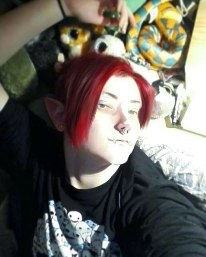 An edited image of a much younger me (white, nb, with bright red short hair and green eyes) laying in bed with plushies and a hand held up, it's made to resemble my OC CK (same as me, but male and pointy ears + cat-like pupils).